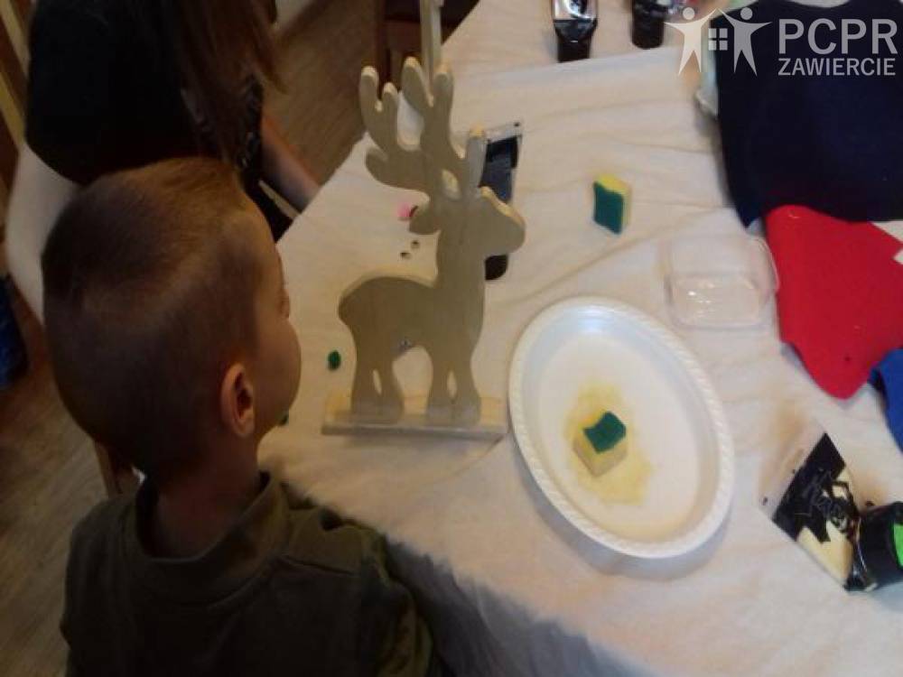 Zdjęcie: Chłopczyk siedzący przy stole, na którym stoi drewniana figurka renifera i rozłożone są tubki z farbami, gąbki do nakładania farb